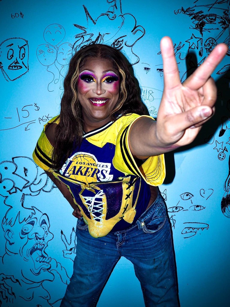 A drag queen in purple makeup and a yellow Lakers shirt