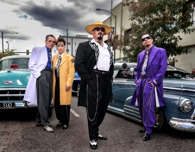 People dressed in zoot suits stand near low-rider cars