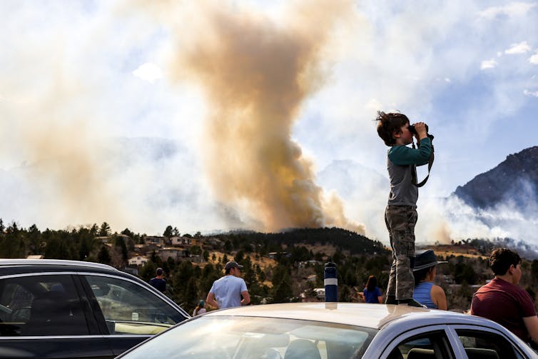 A boy stands on top of a car, peering through binoculars, as orange smoke billows in the background.