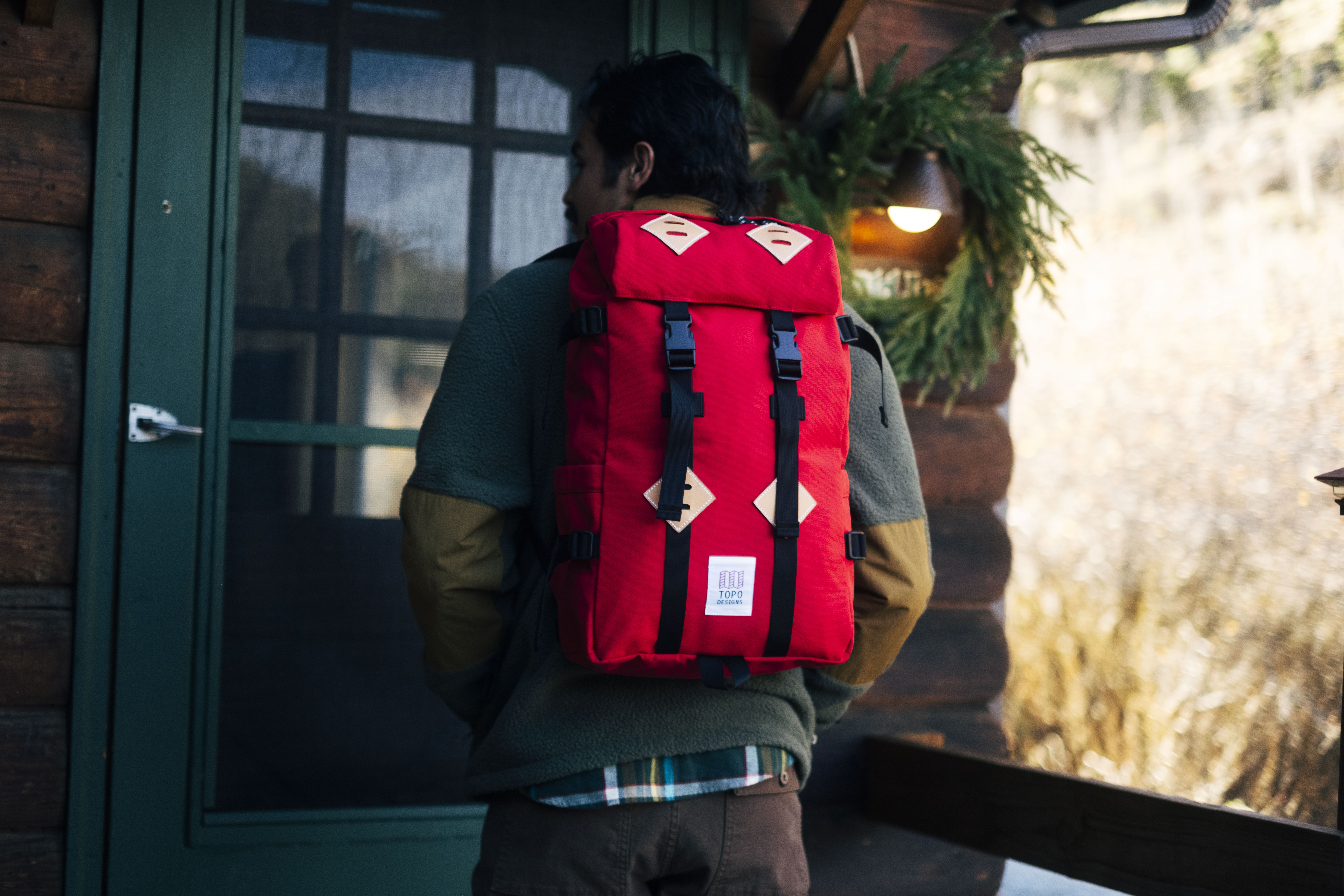 A person wearing a red Topo Designs backpack and apparel outside of a log cabin