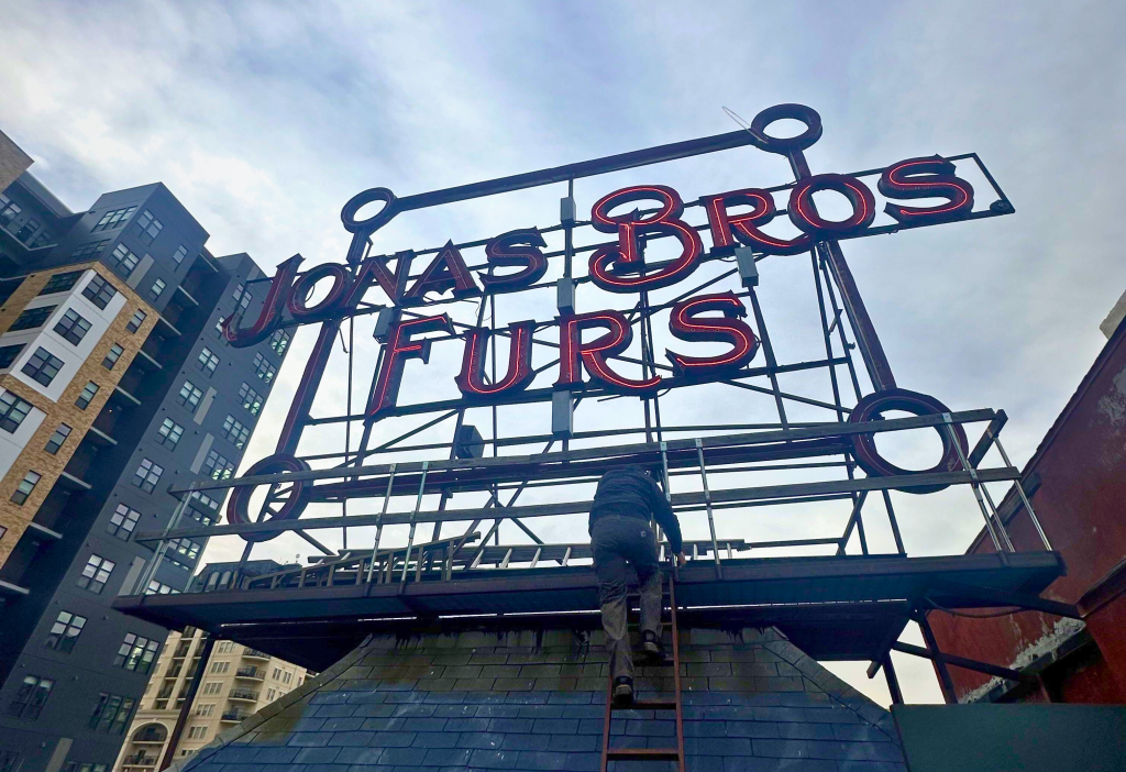 A man climbs a ladder up to the red "Jonas Bros Furs" neon sign atop a roof.