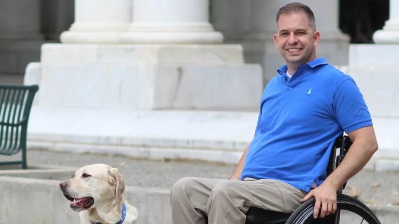 A man sits in a wheelchair to the right of a golden retriever.