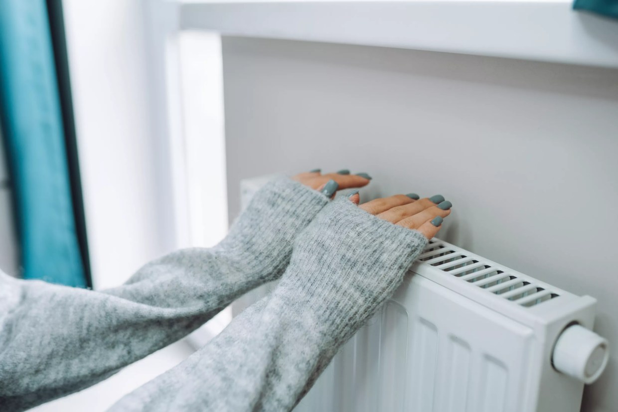 Someone wearing a grey sweater warms their hands on a radiator.