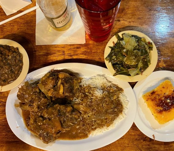 a beer on a table with a plate of oxtails and rice and a bowl of collard greens on the side.