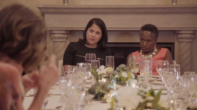 Women sit at a fancy table covered in wine glasses and white flowers.