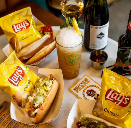 several hot dogs on a table along with a cocktail and a bottle of wine