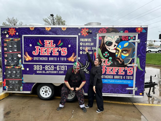 a man in a lucha libre mask and a woman stand in front of a food truck.
