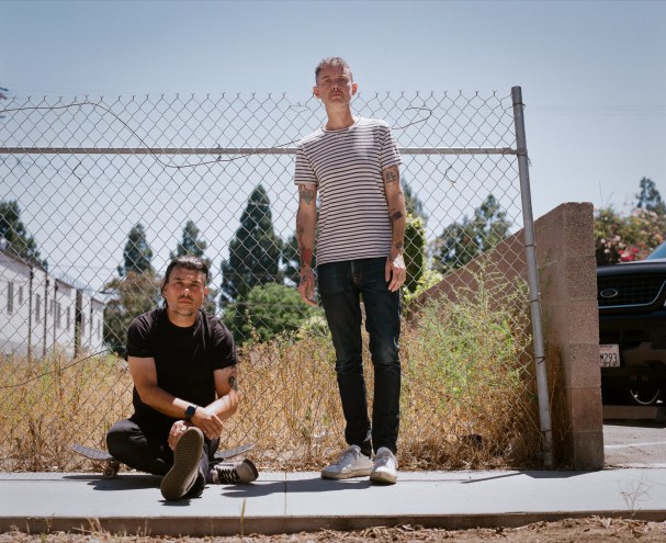 two men pose in front of a chainlink fence