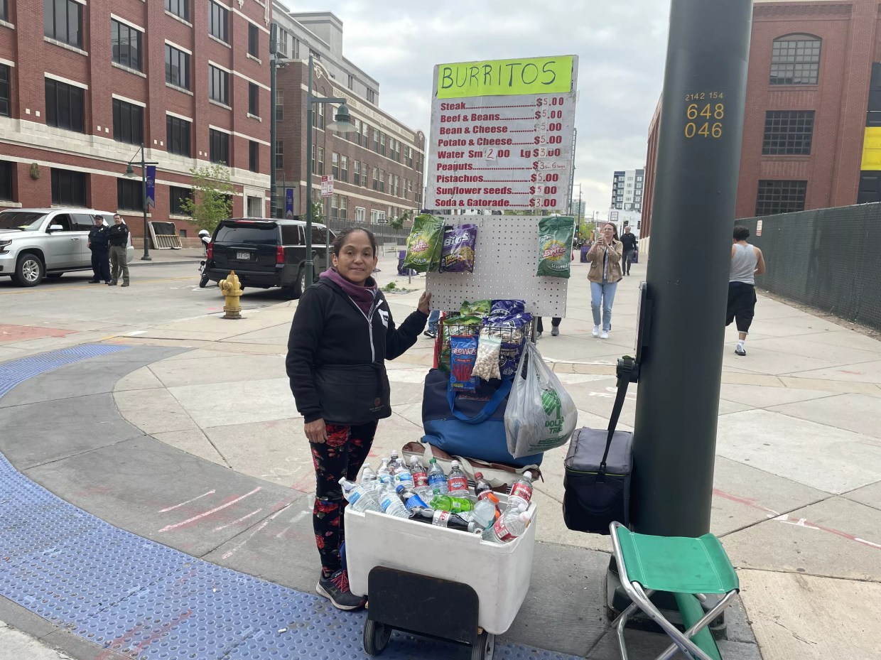 A woman stands on the corner with a cart containing ballpark snacks.