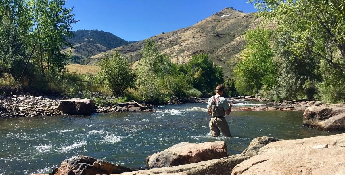 A man wearing fishing waders casts a pole into a clear river.
