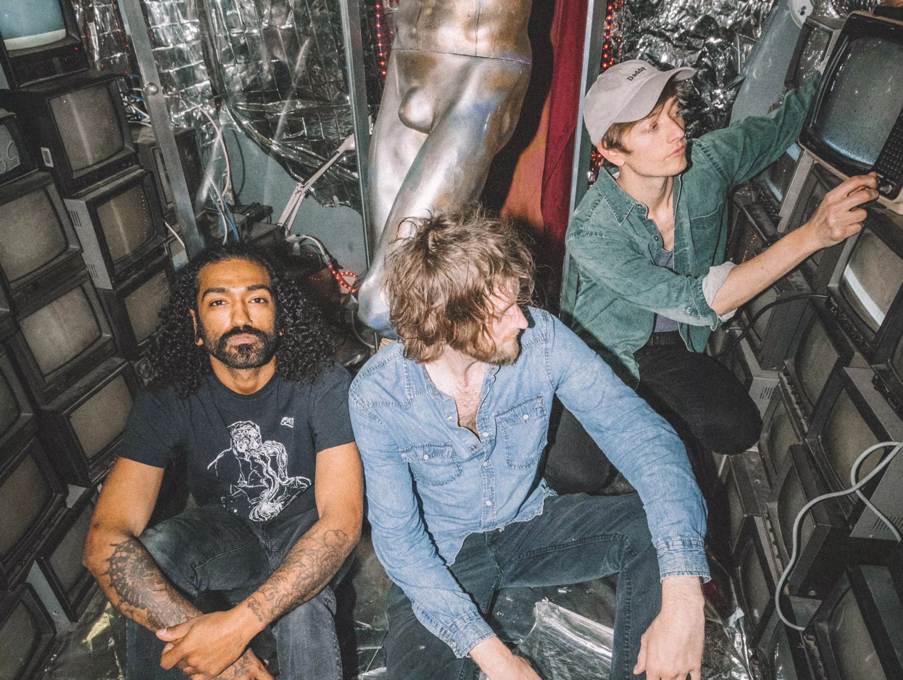 Three men sitting and squatting in front of a statue of legs, surrounded by vintage box TVs.