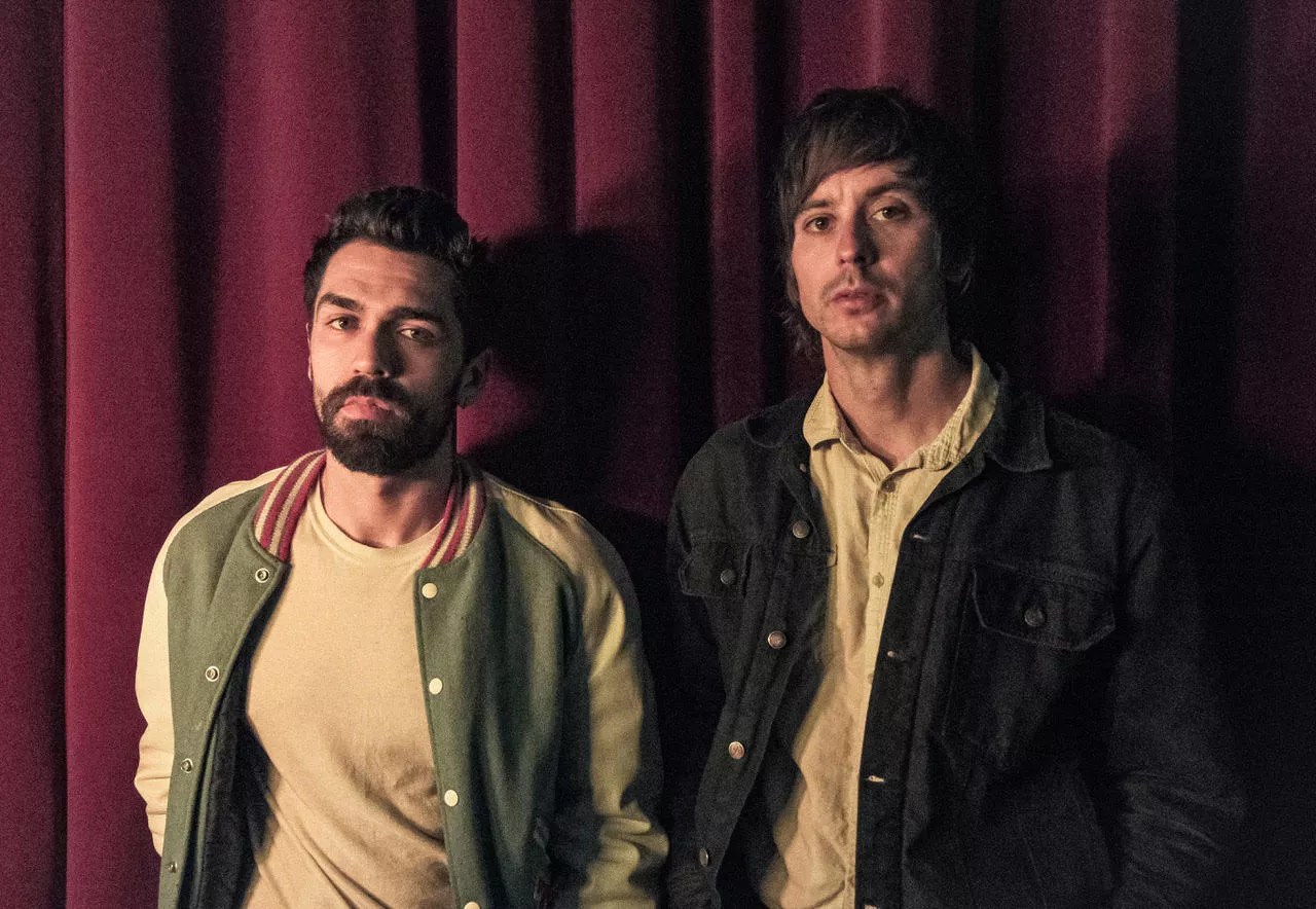 two men in jackets stand in front of a red velvet curtain.