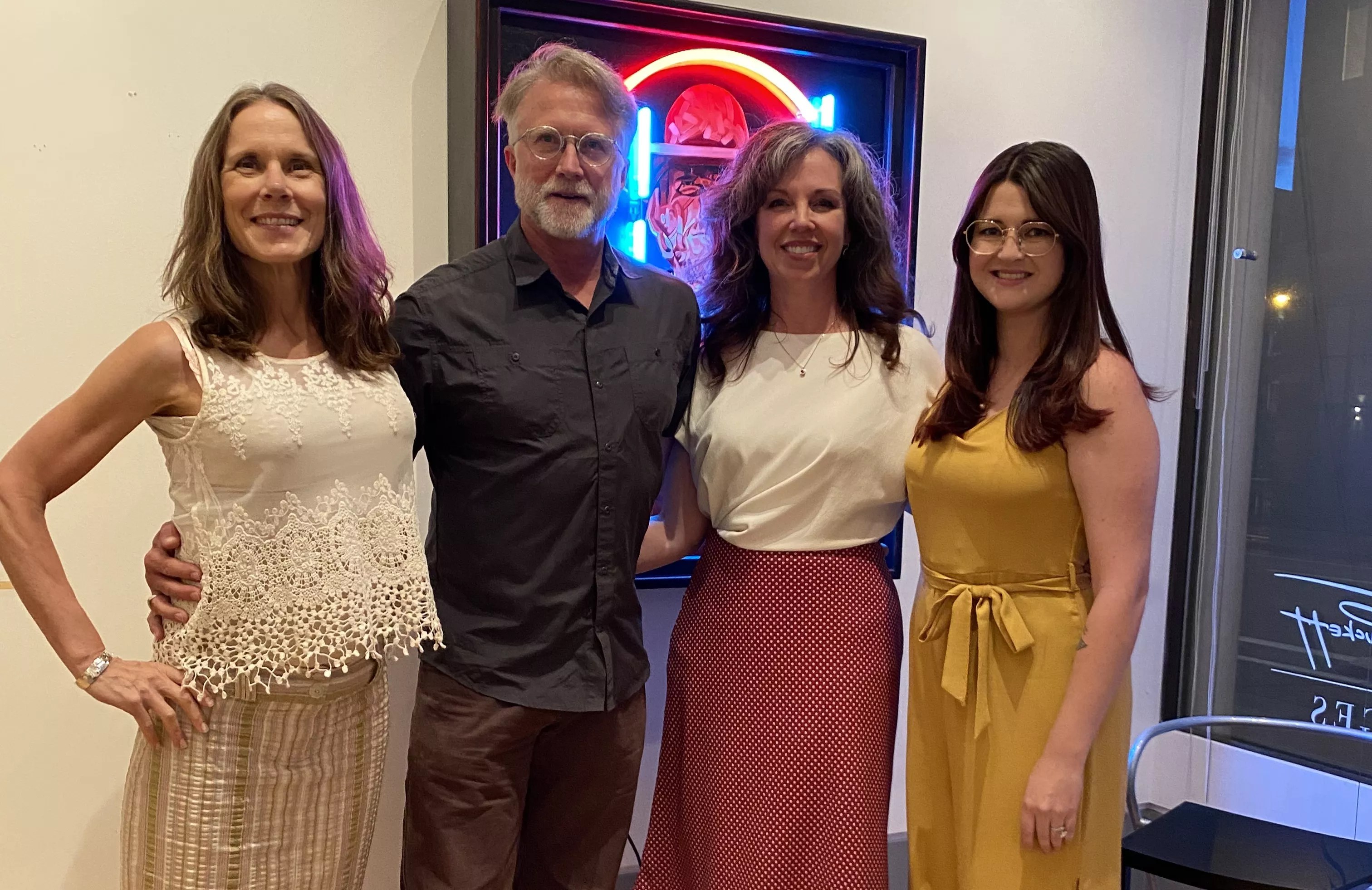 woman in white shirt, man in navy shirt, woman in white shirt and skirt, and woman in yellow post in front of a painting.