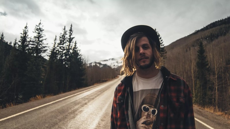 A man with long blonde hair standing next to a road with trees on left.