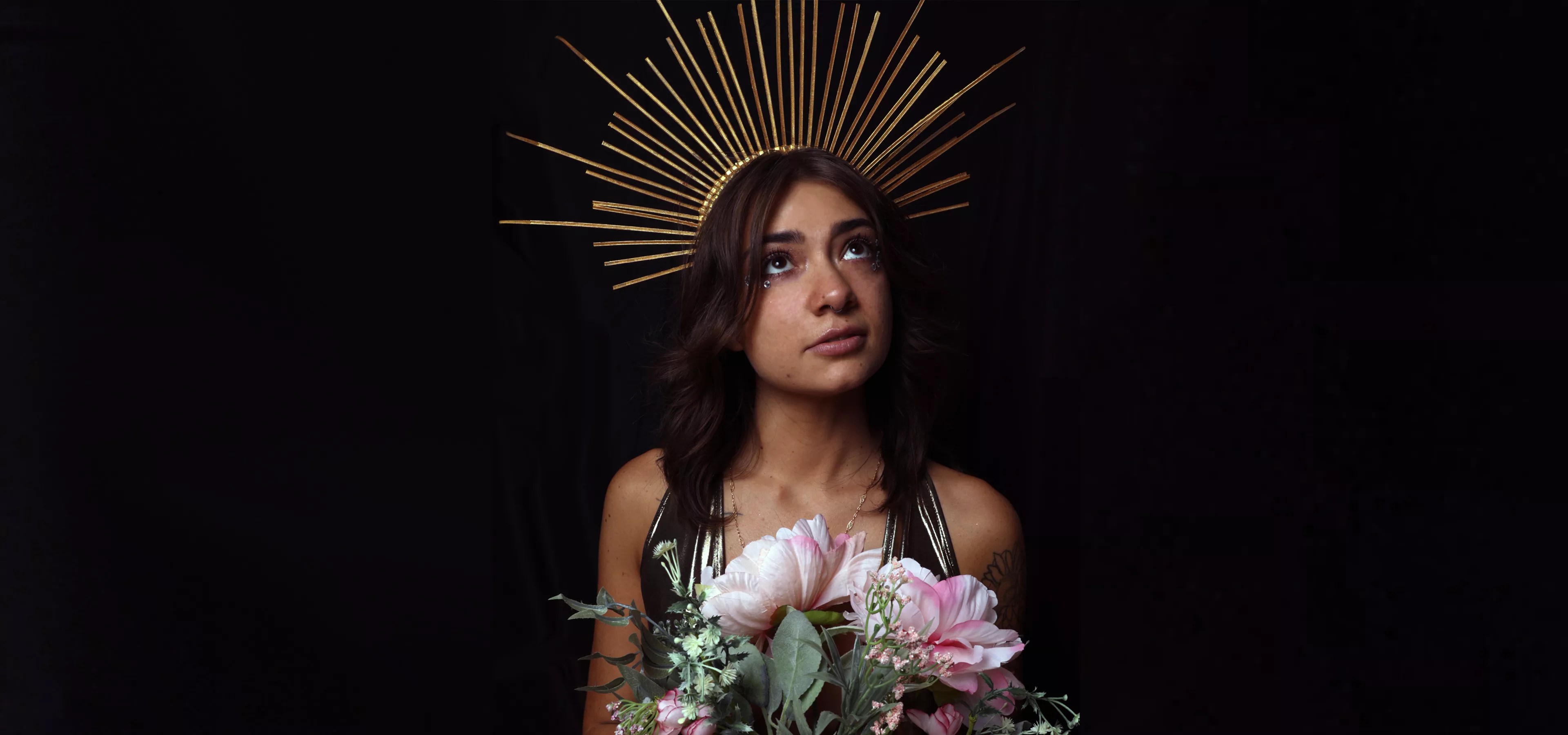 brunette woman wearing gold crown and holding pink flowers.