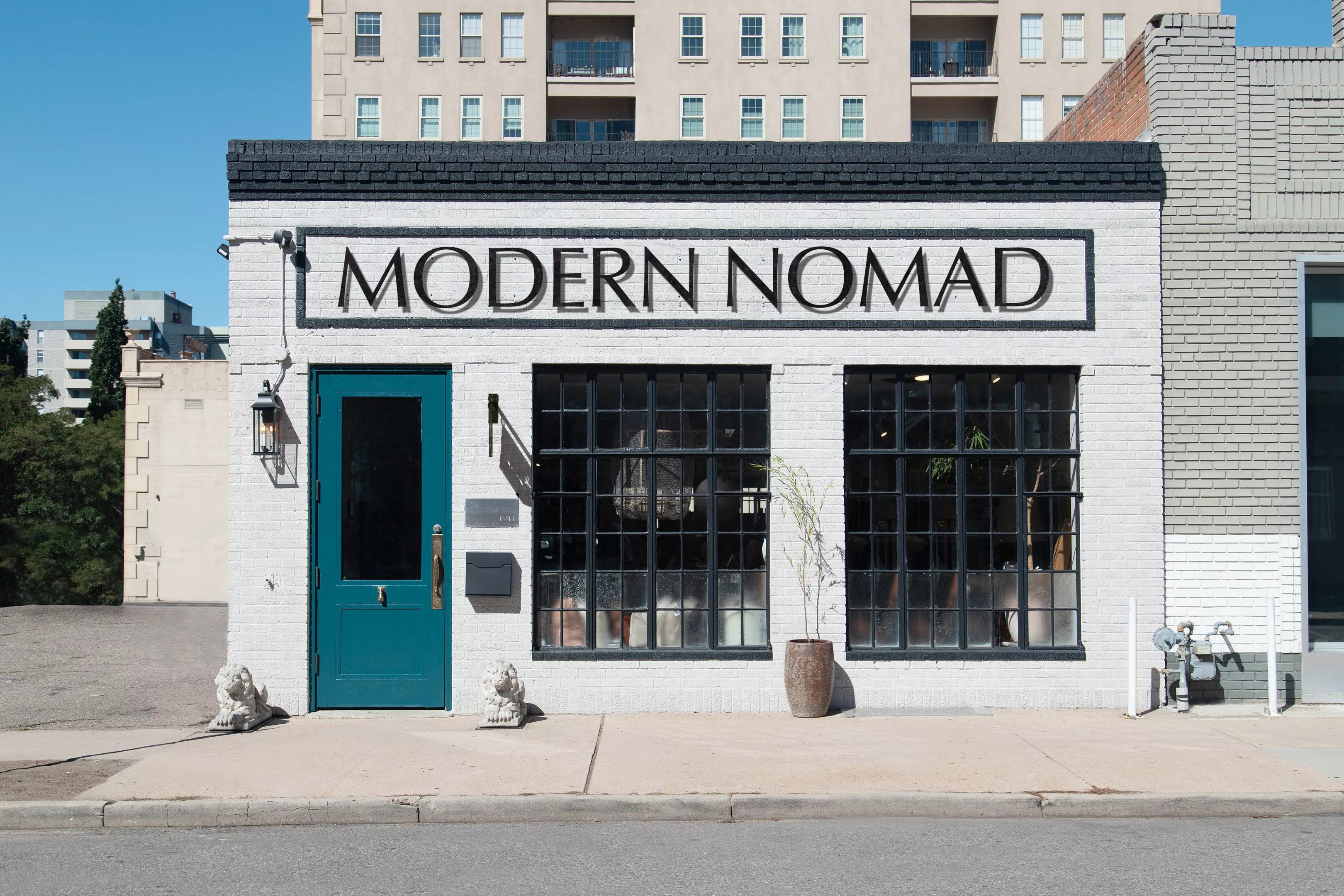 The front-facing exterior of the Modern Nomad. Square building with white walls, two large windows and a cyan door. Name in large capital lettering near the roof.