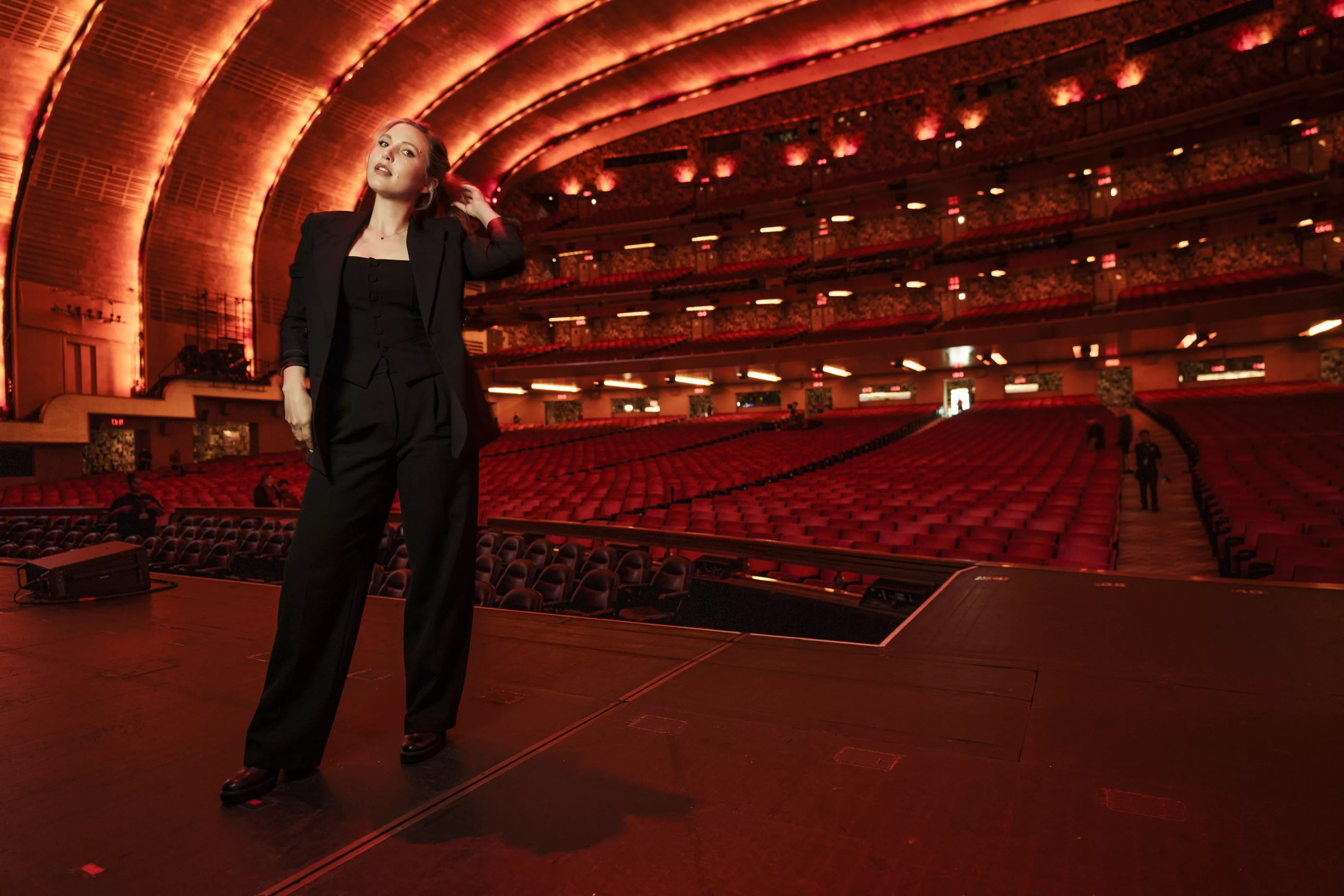 comedian taylor tomlinson stands in a theater in a black pant suit.
