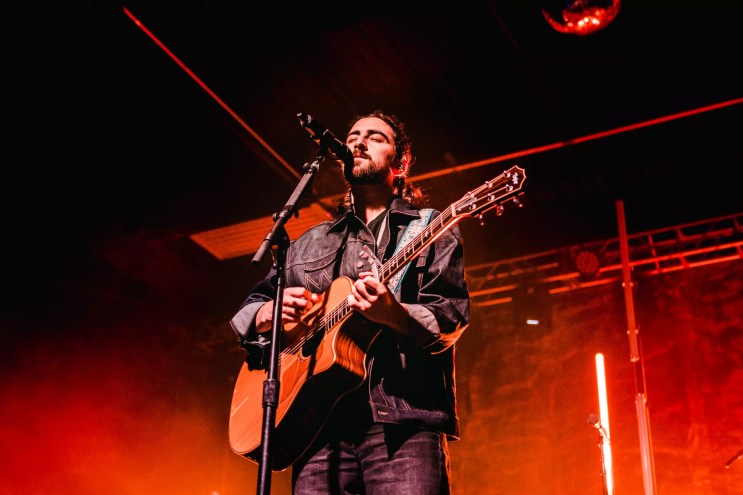 man with guitar singing into a microphone against a red background