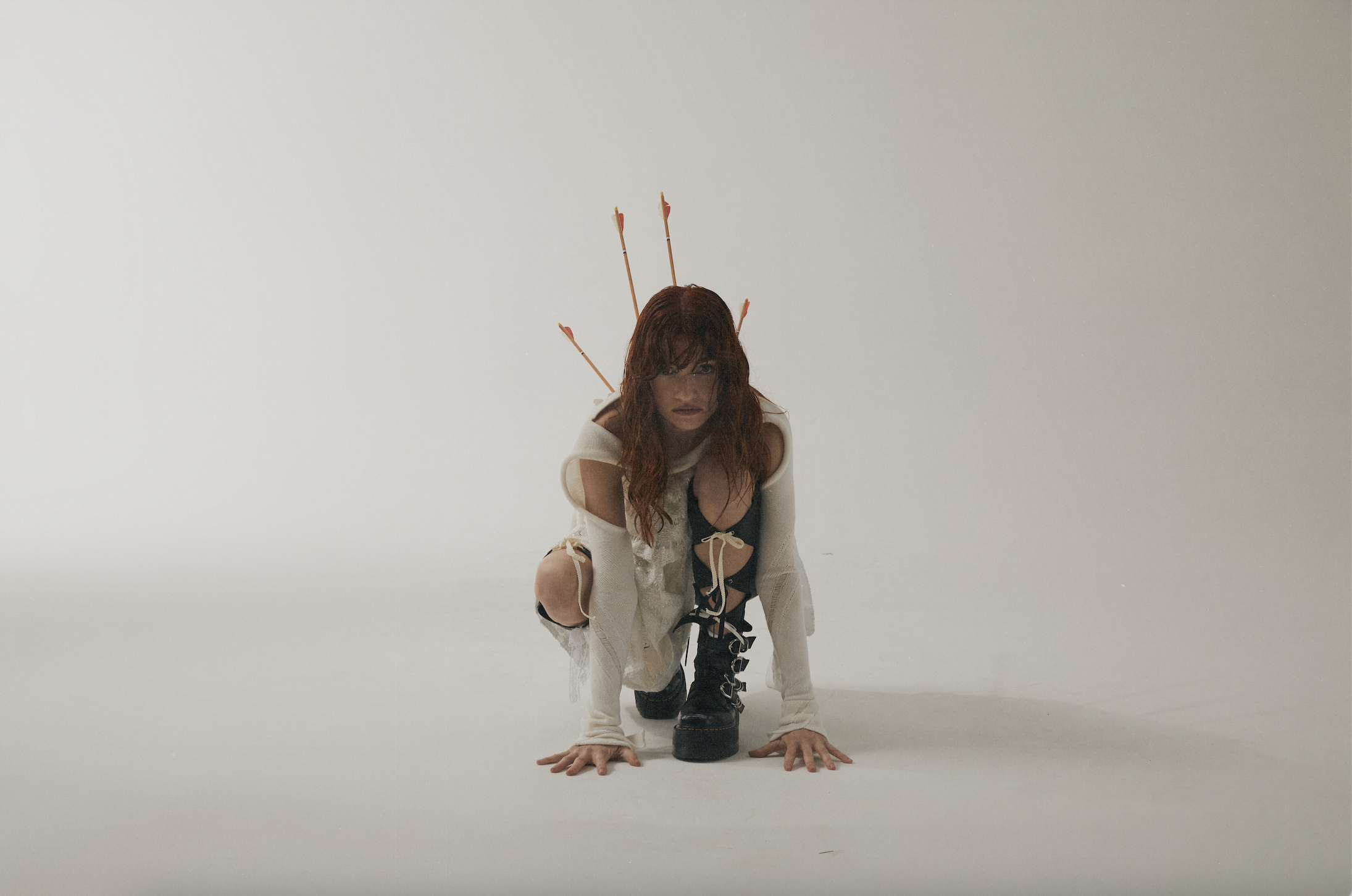 woman crouches in white dress with arrows sticking out of her back