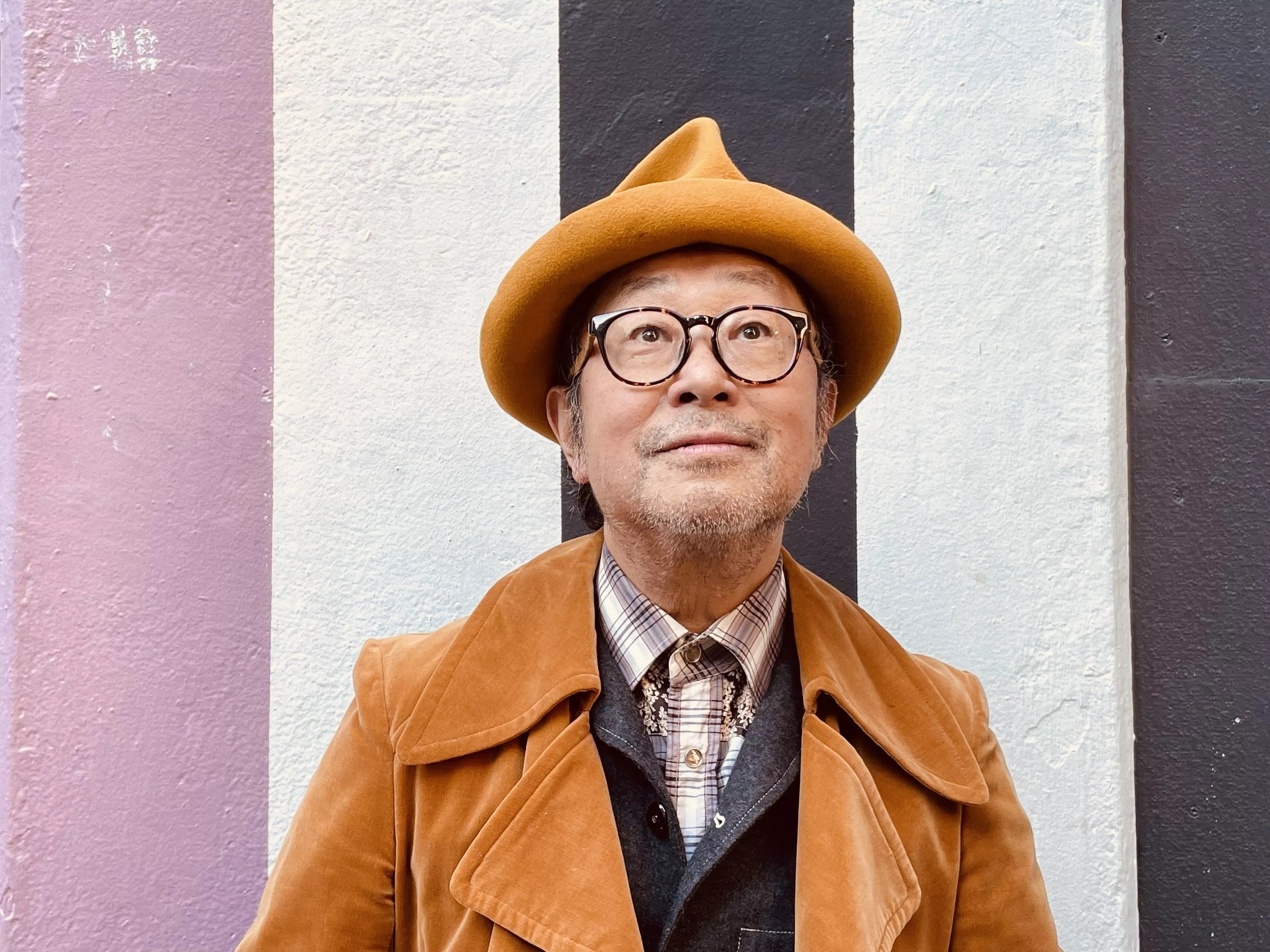 Portrait of Senri Oe in brown hat and Jacket on vertically striped background.