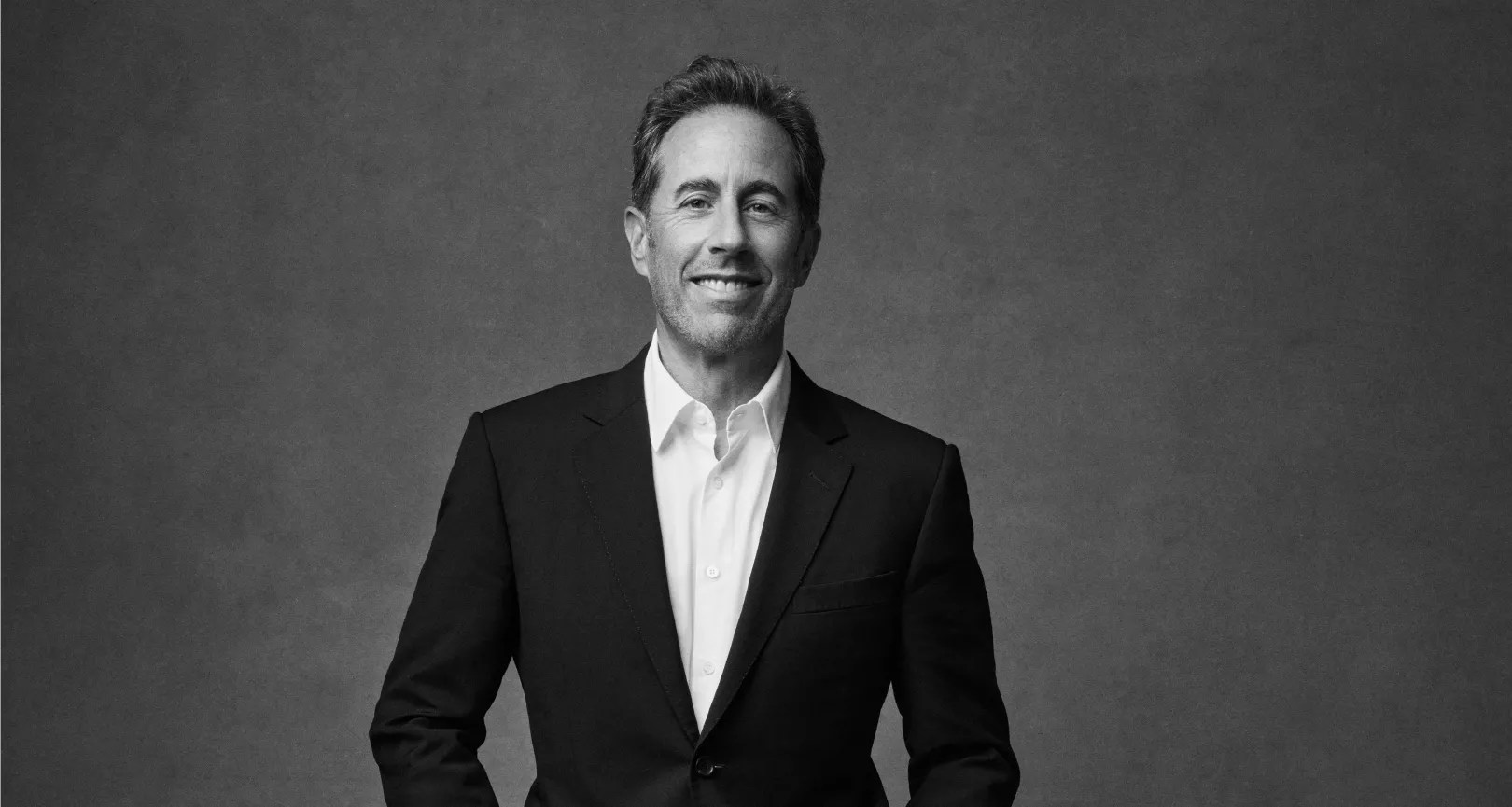 jerry seinfeld poses in a black suit and white shirt