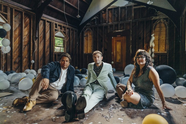 two men in suits and a woman sit on a floor in a wood panel room