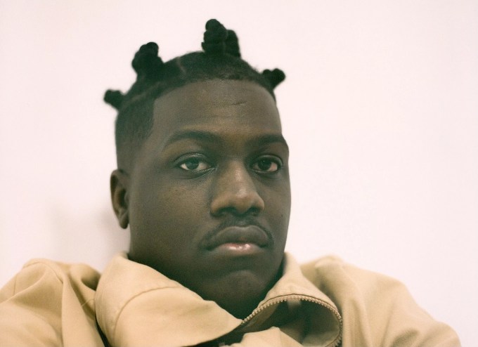 black man in an orange sweater sitting against a pink wall