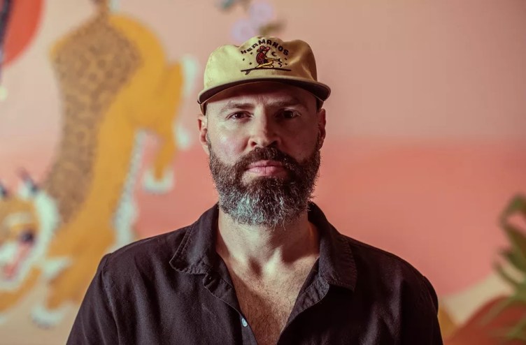 man with beard in a flat brim hat and button down shirt in front of a pink background