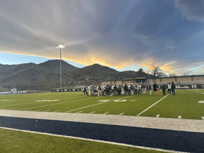 Under a cloudy sky, a football team huddles on a green turf field.