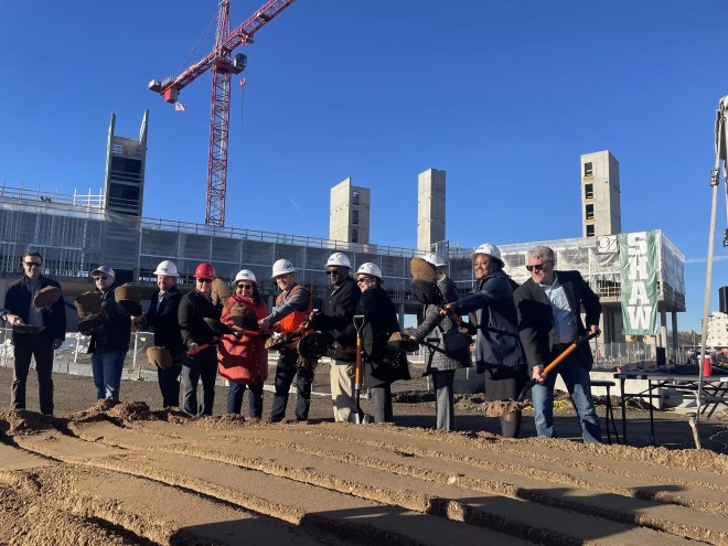 A line of people wearing white hard hats toss dirt in the air using orange-handled shovels.