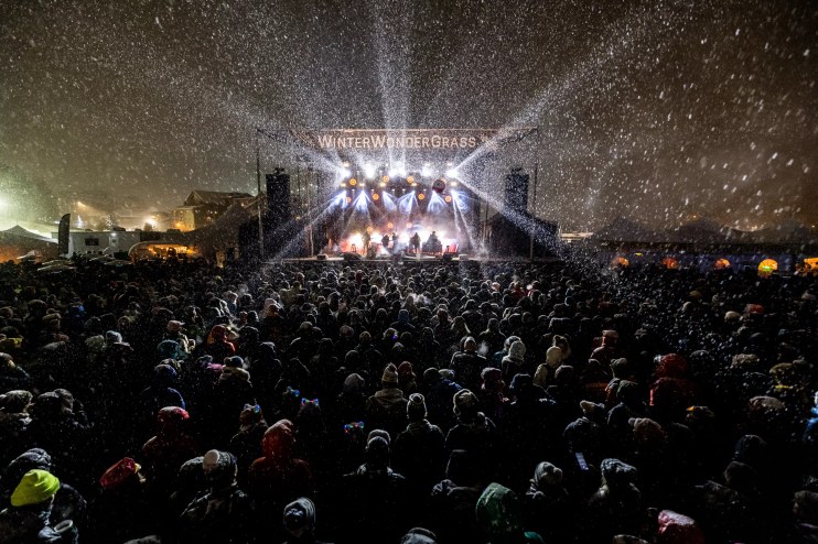 a stage with the words WinterWonderGrass is surrounded by an audience in the snow