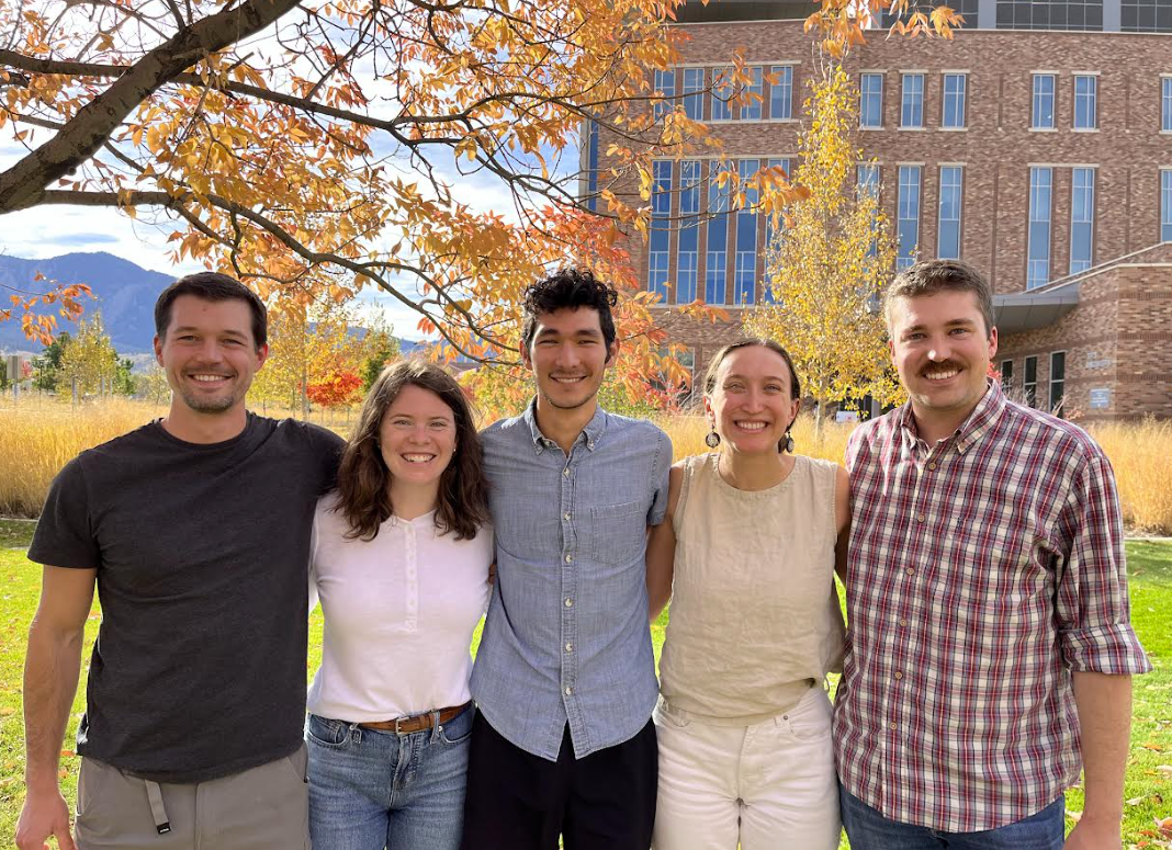 Five people smile at the camera arm in arm.