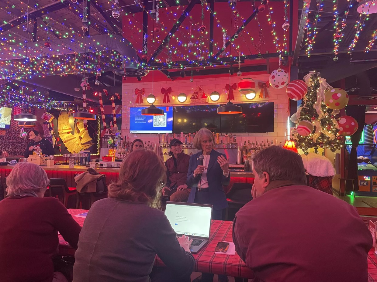 A woman in a blue suit speaks to people gathered in a room decorated for Christmas.