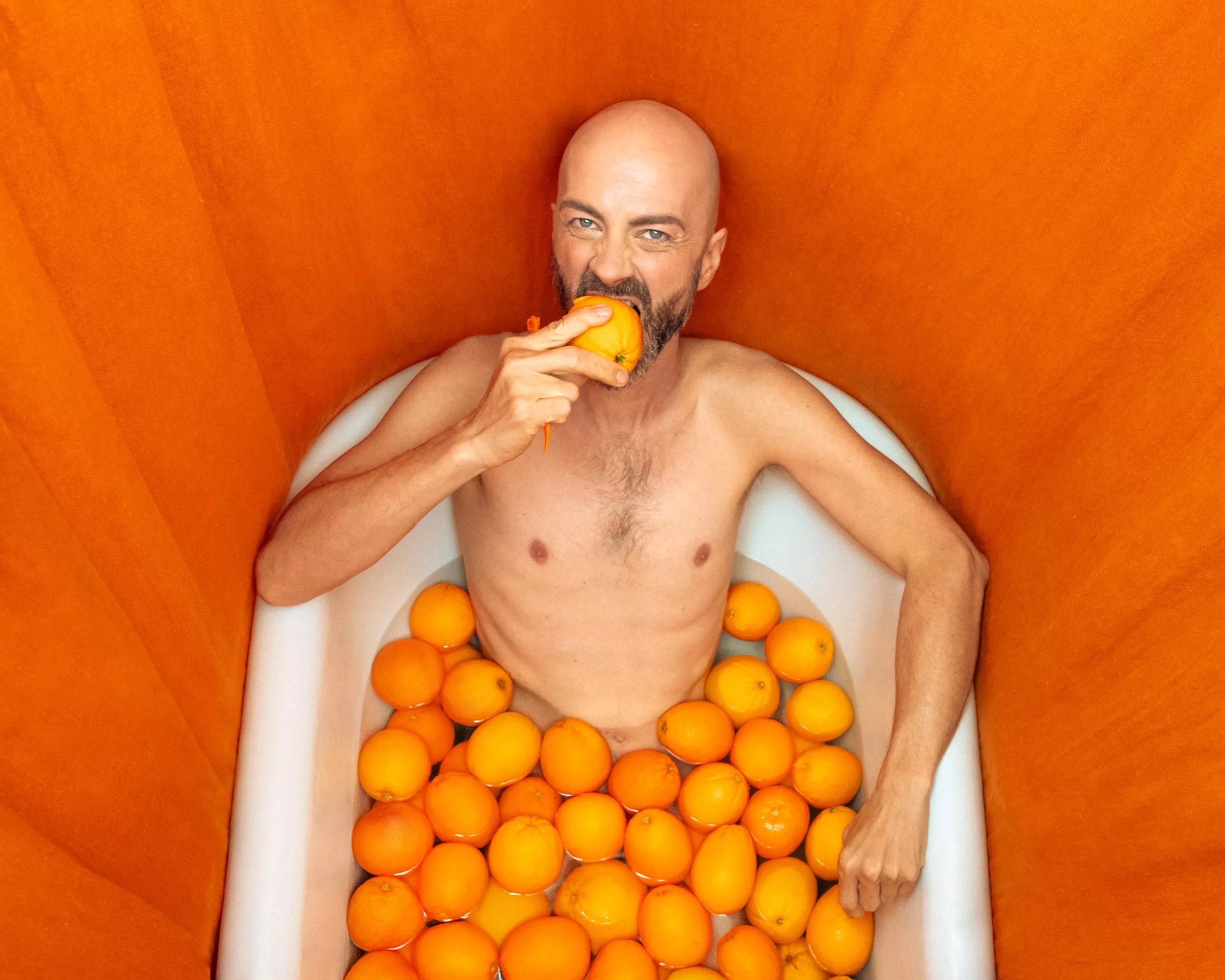 man eating an orange in a bathtub filled with oranges