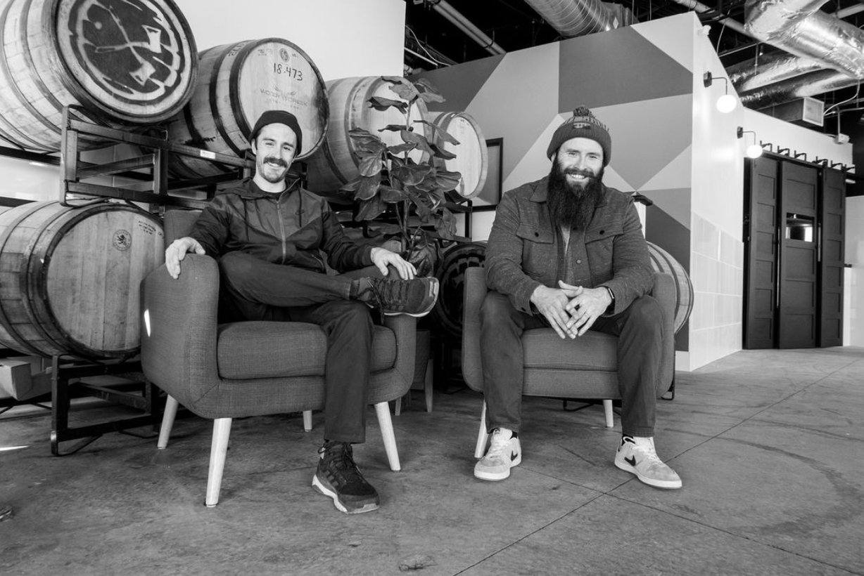 Two men sitting in front of beer barrels.