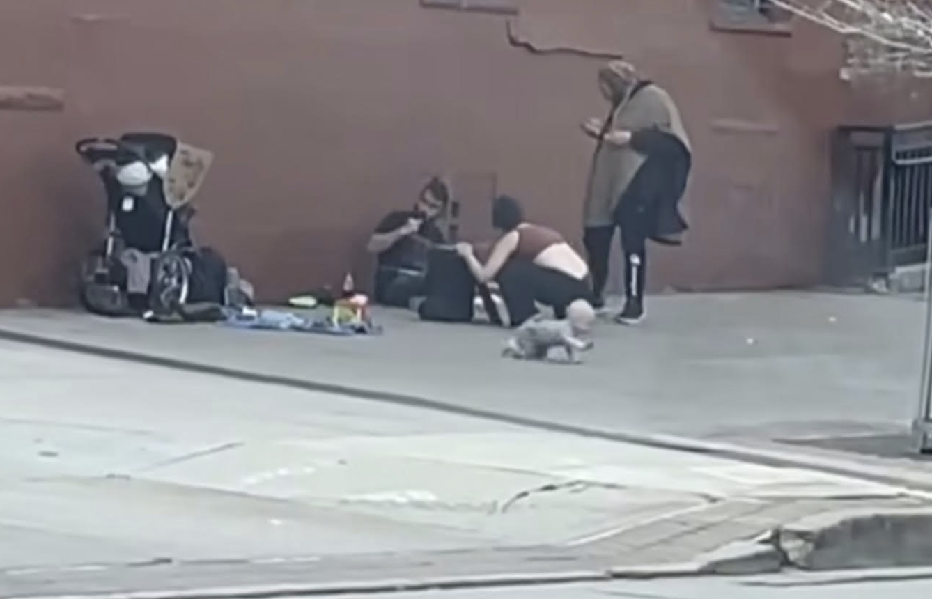 A baby crawling on a sidewalk while his mother and friends hang out nearby.