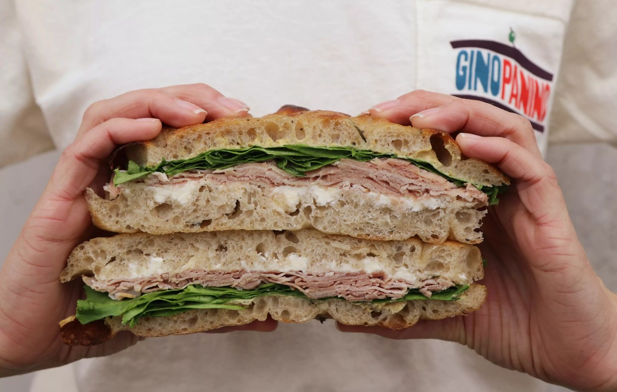 A person in a branded Gino Panino shirt holding a ham and mascarpone sandwich