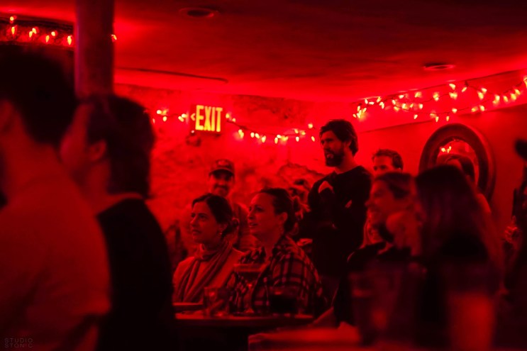 The audience sits and stands in a room lit with red lights.