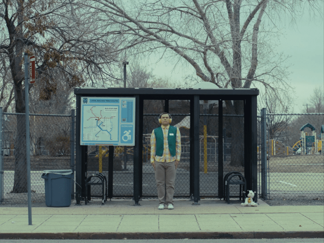 Man with headphones stands at a bus stop.