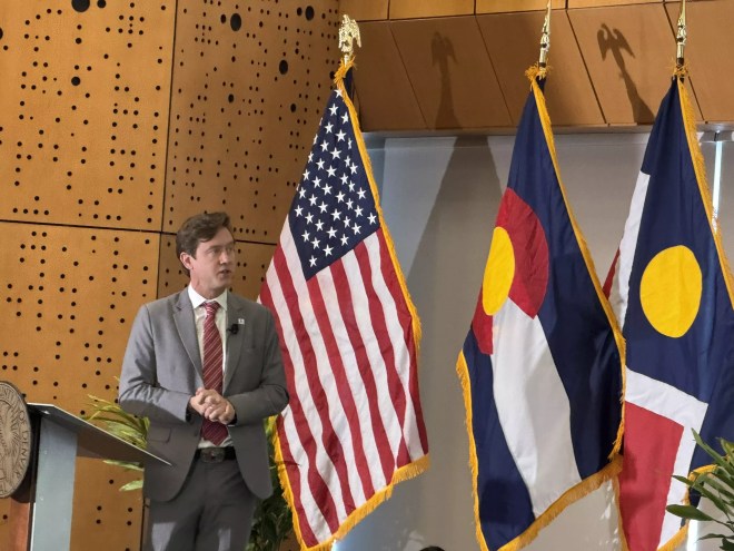 Man in a suit speaks in front of American and Colorado flags