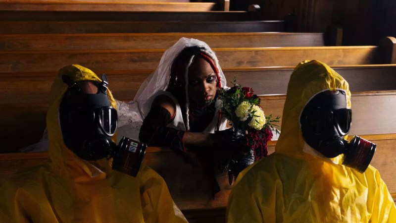 two people in hazmat suits sit around a bride in church pew