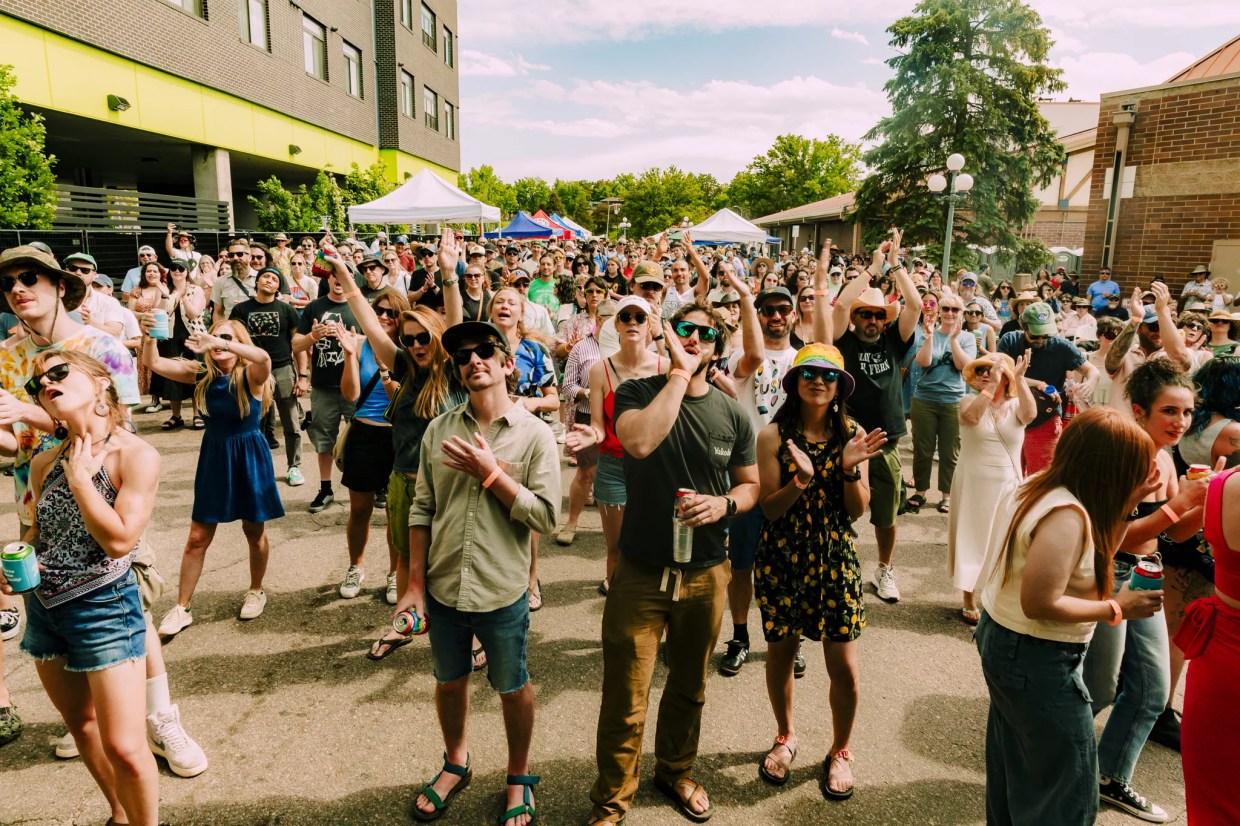 A large crowd of music fans cheer for musicians. Tents with vendors and food trucks are in the distance behind them.