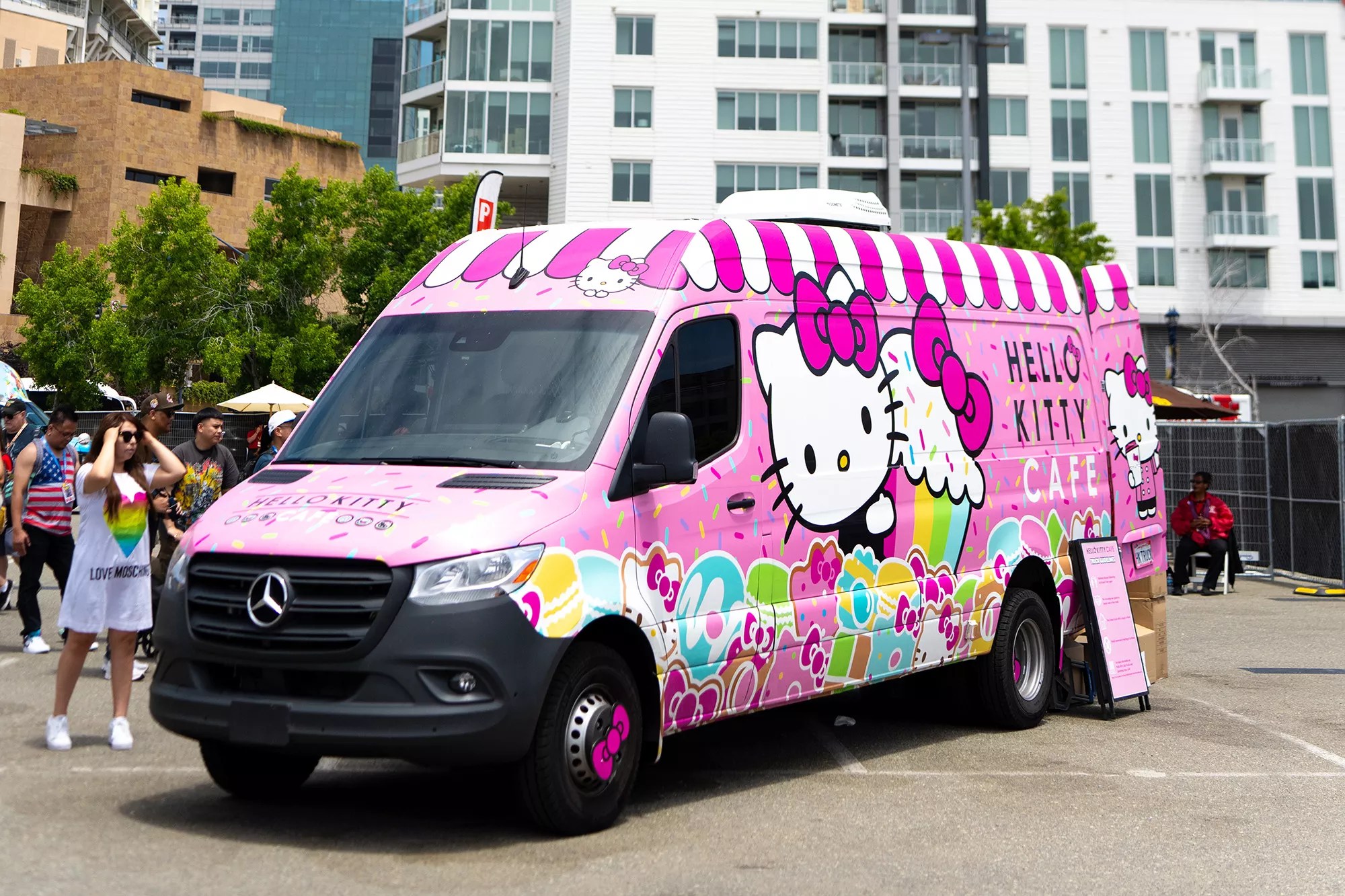 A pink Hello Kitty-themed truck serves sweet treats and drinks to customers on a sunny day.