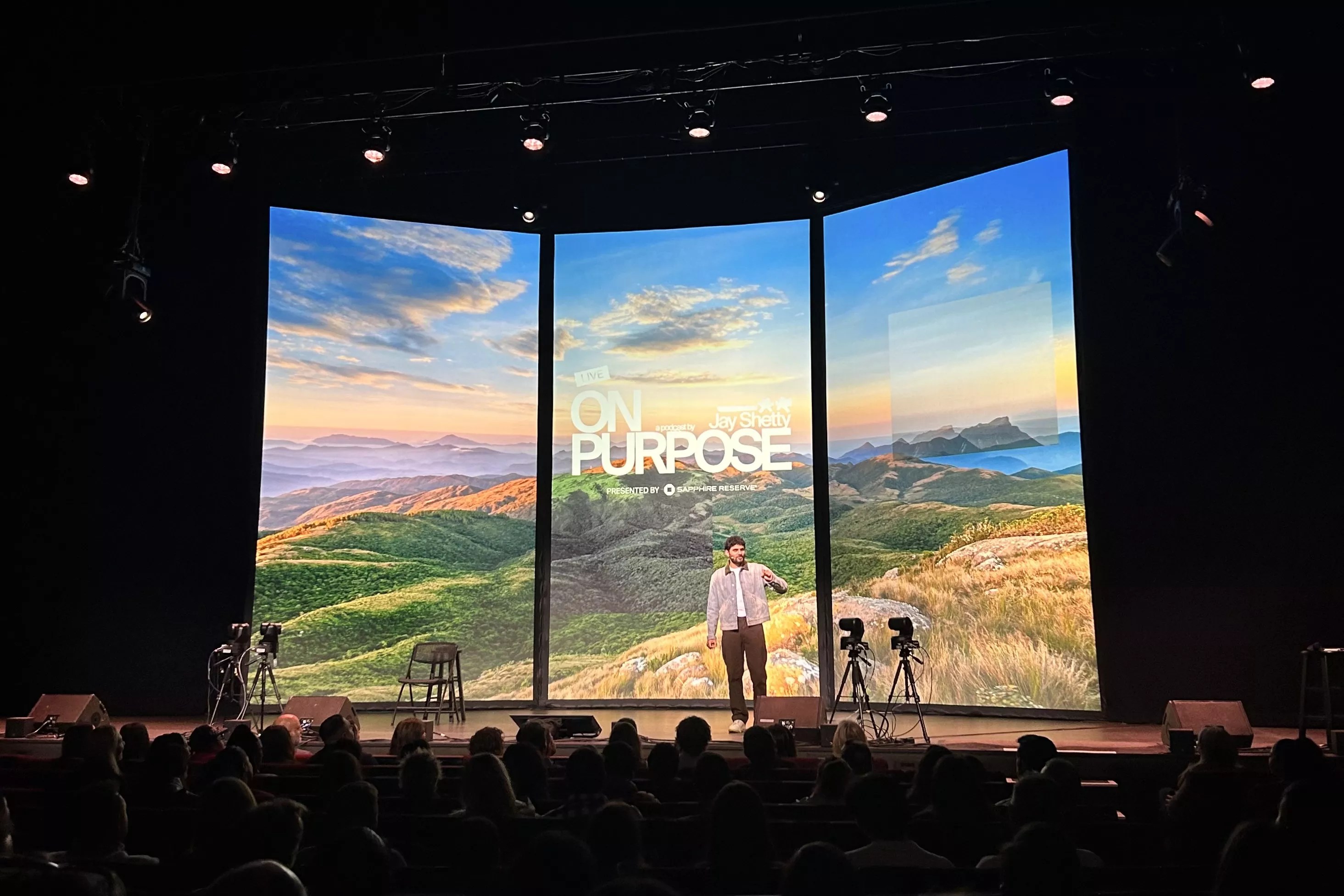 Jay Shetty backdropped by a mountain scene on three digital panels while on stage at the Ellie Caulkins Opera House