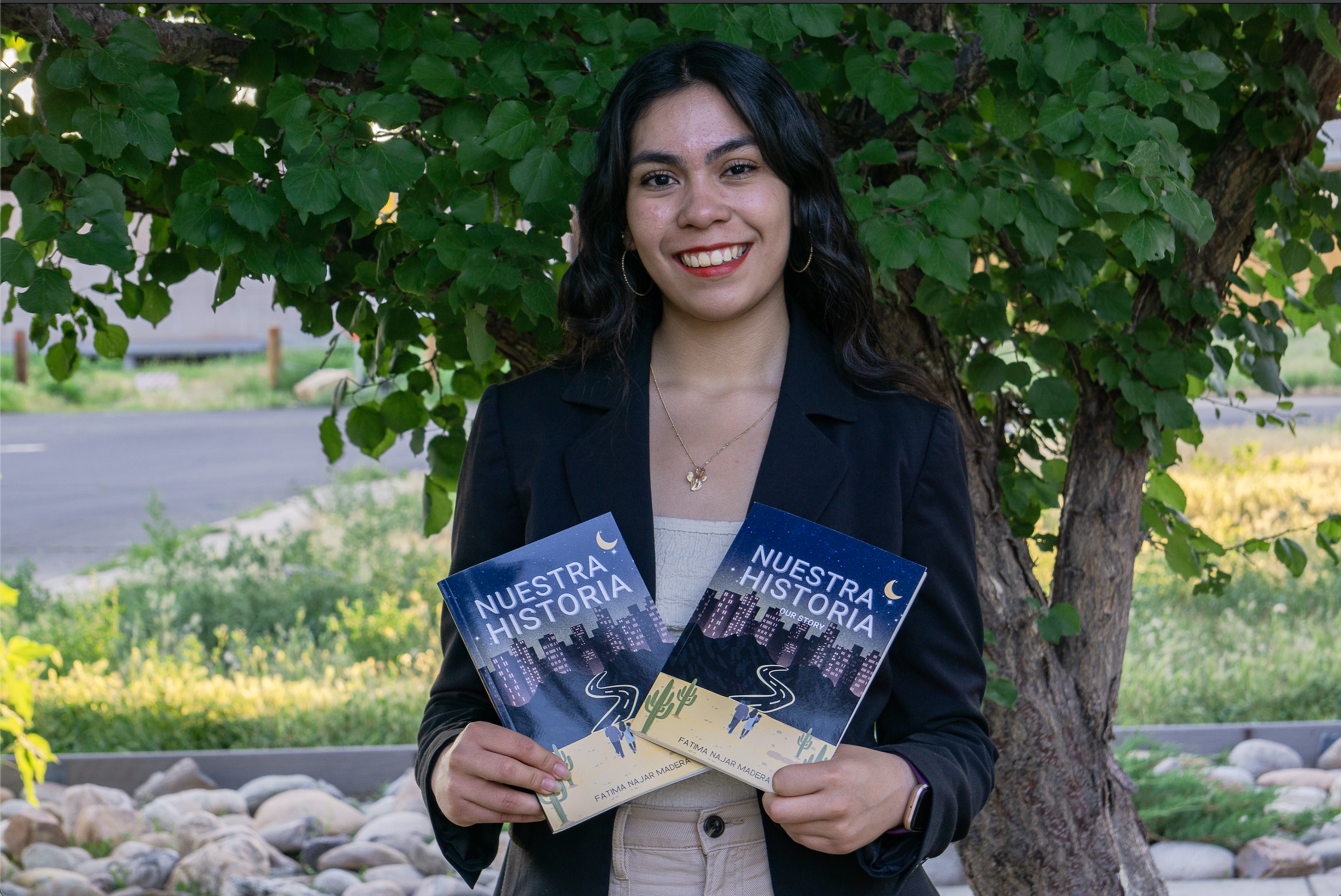 A girl wearing a black blazer, gold hoop earrings and a bold red lip smiles while holding two books in front of greenery.
