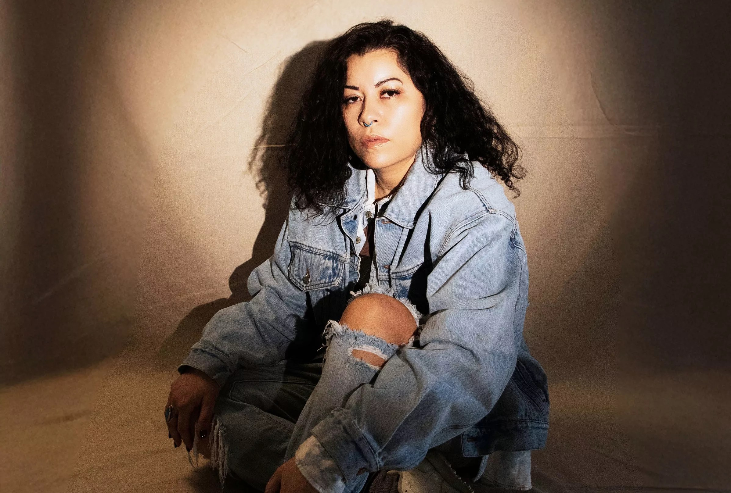 A woman wearing a all-denim look and turquoise nose-ring poses in front of a beige backdrop for the camera.