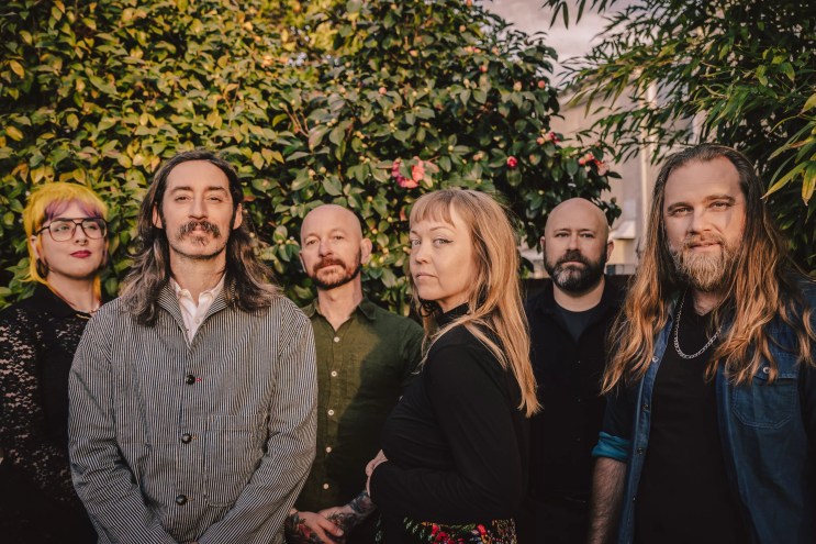 The six current members of indie rock band Murder By Death stand under some greenery.