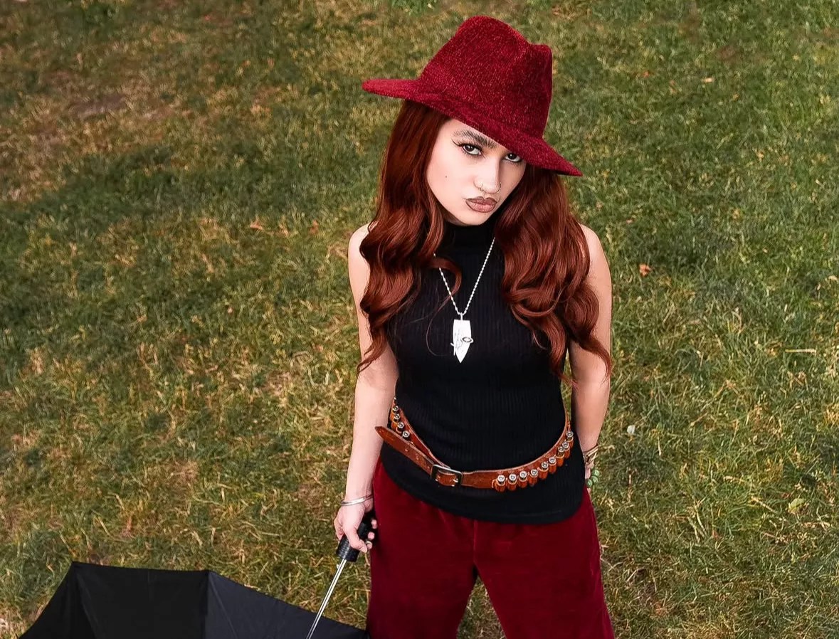 Kitty wears a red suede hat and pats with a black top while holding an umbrella on a lawn.
