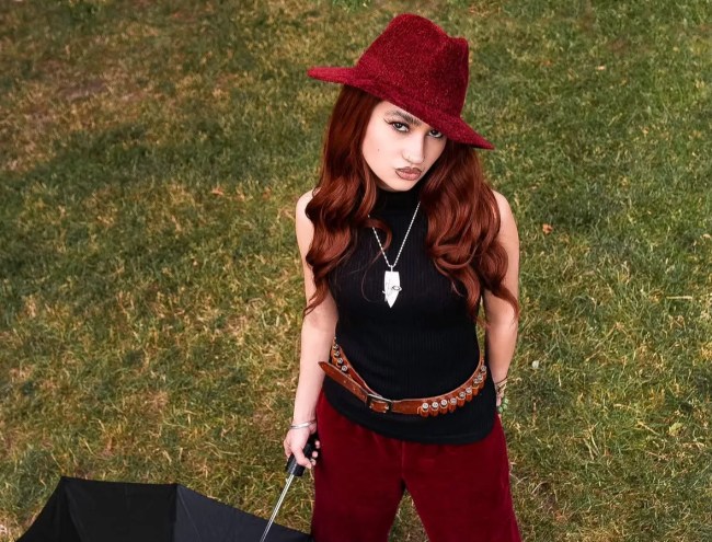 Kitty wears a red suede hat and pats with a black top while holding an umbrella on a lawn.